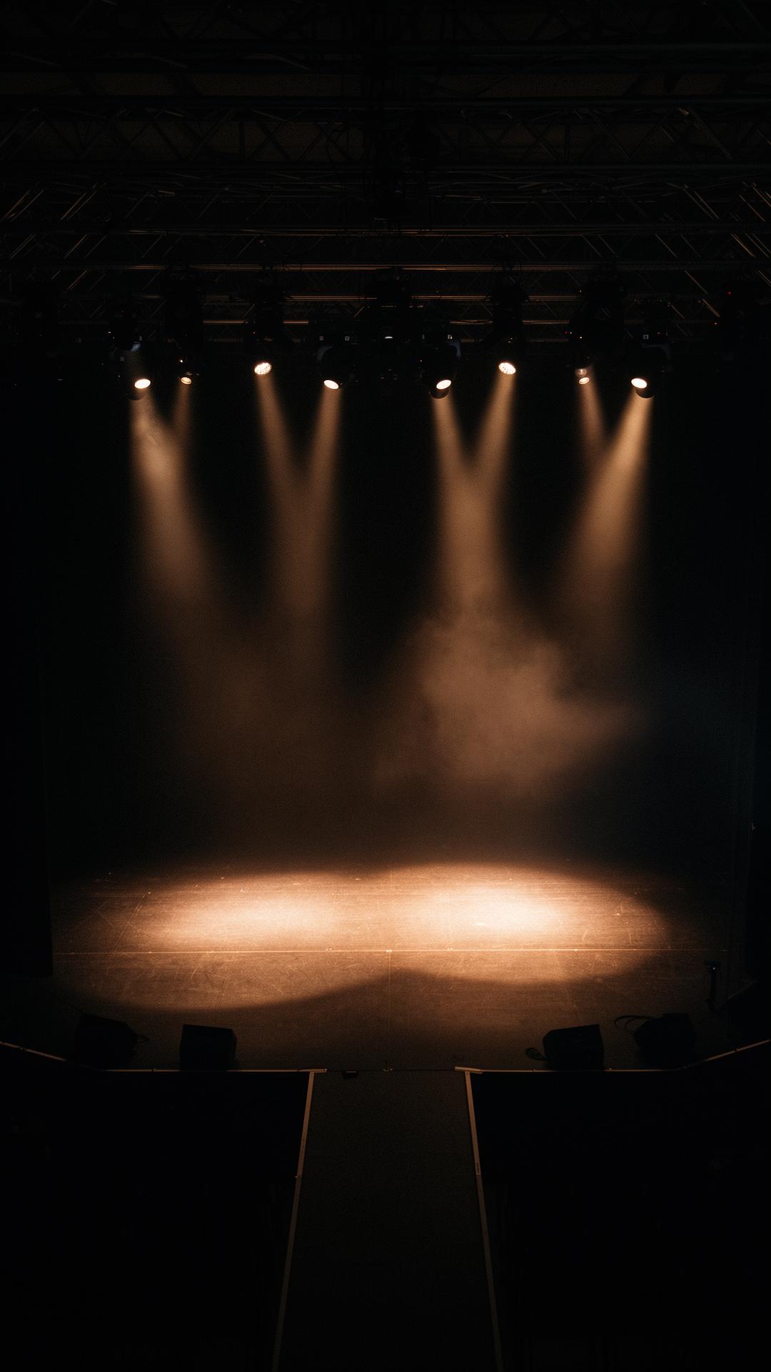 A darkened event stage lit by a row of warm overhead spotlights cutting through atmospheric haze